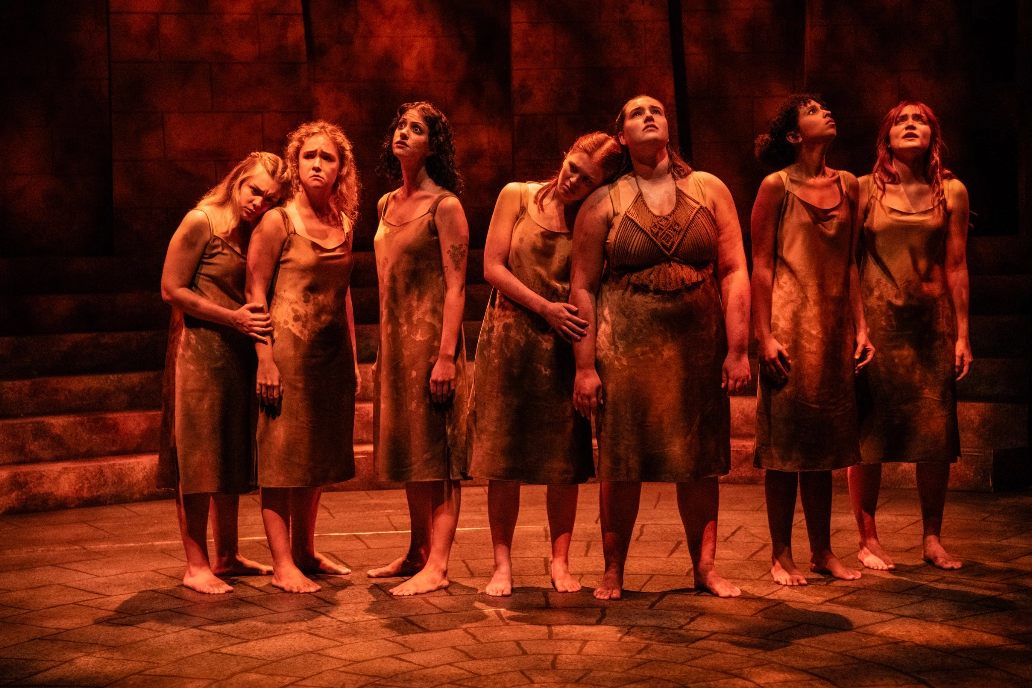 Seven Antigone performers in earth-toned costumes under warm red stage lighting.