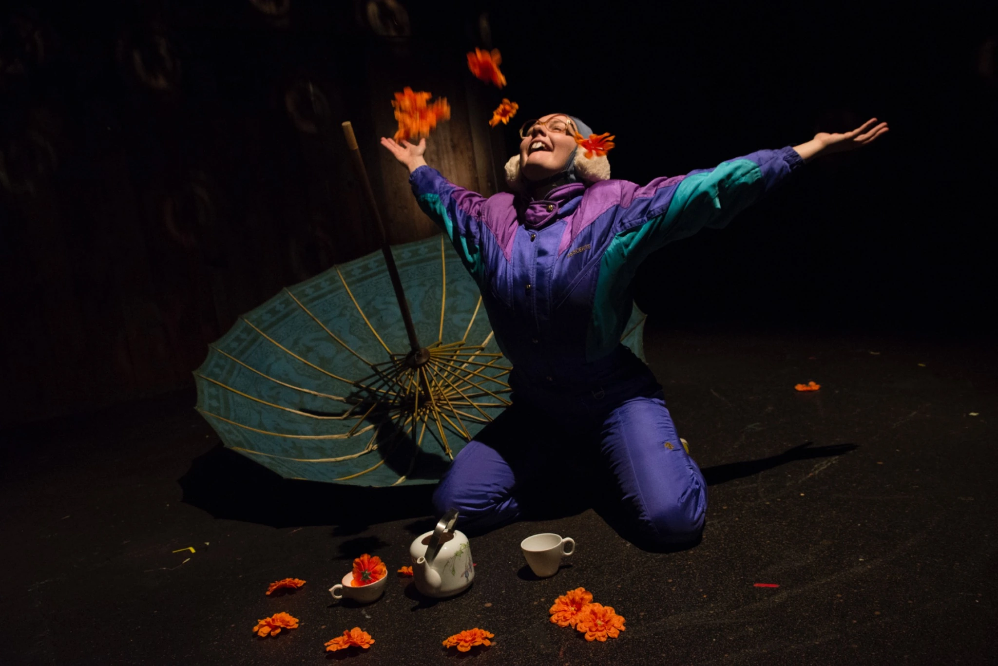 A performer in a purple and teal snowsuit tosses orange flowers beside an open umbrella and tea set on stage.