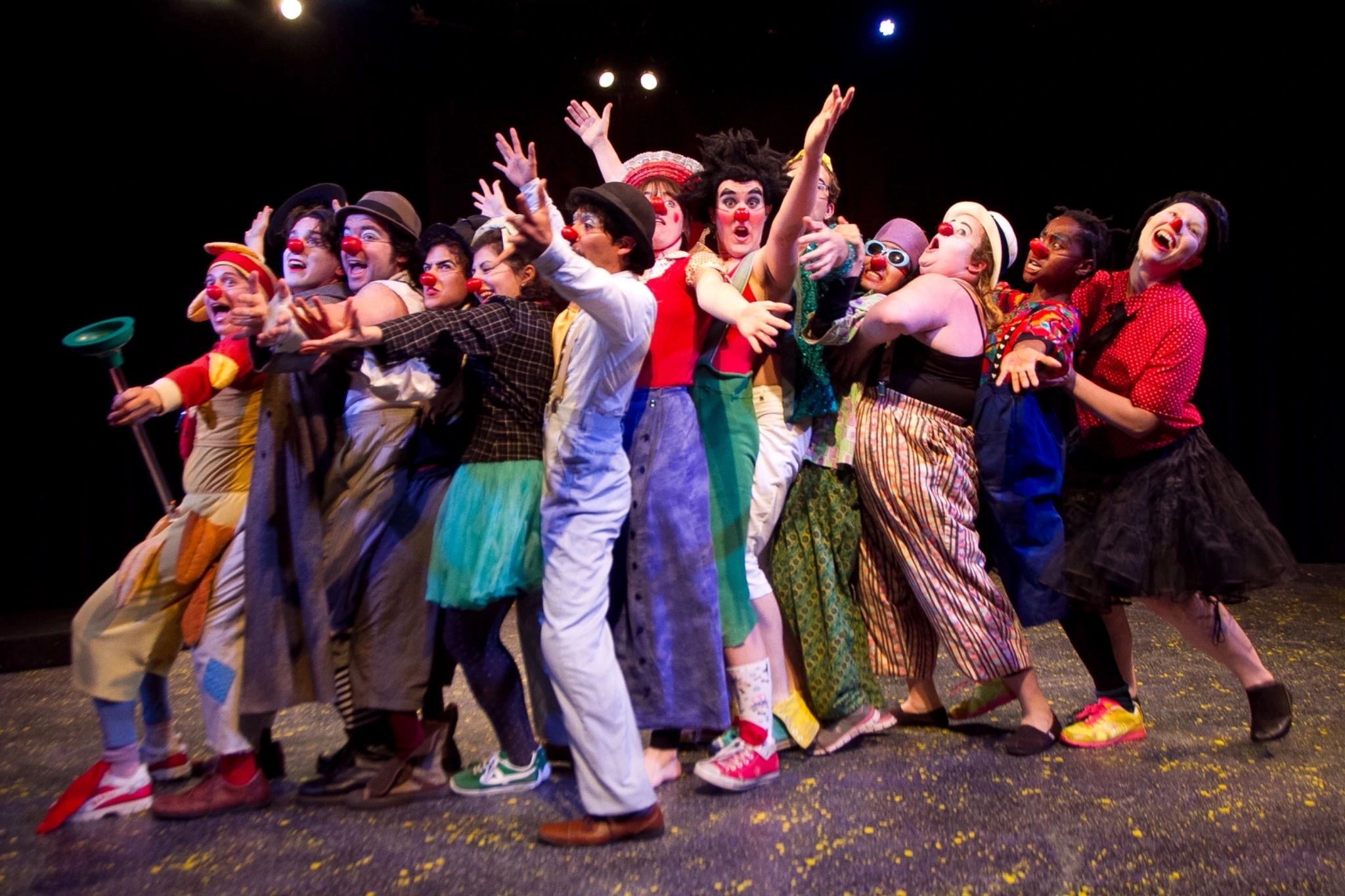 An ensemble of clowns in colorful costumes and red noses reaches outward together on stage.