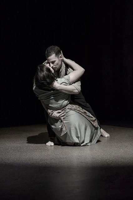 Two performers kneel together in an embrace on a dark stage.