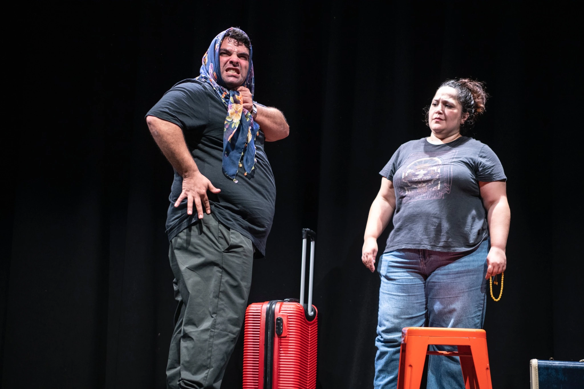 Two Kismet performers on a dark stage beside a red suitcase and an orange stool.